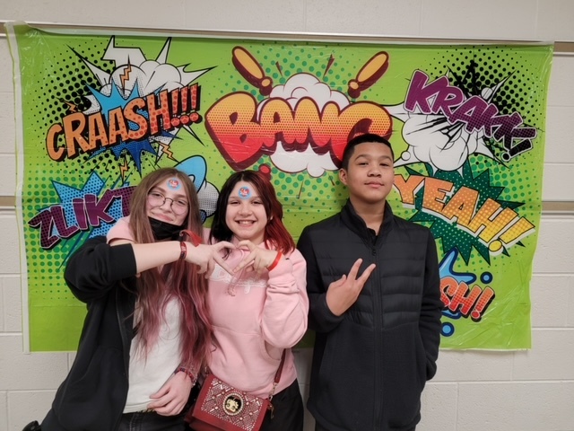 Three middle school students smile for a photo in front of comic book backdrop